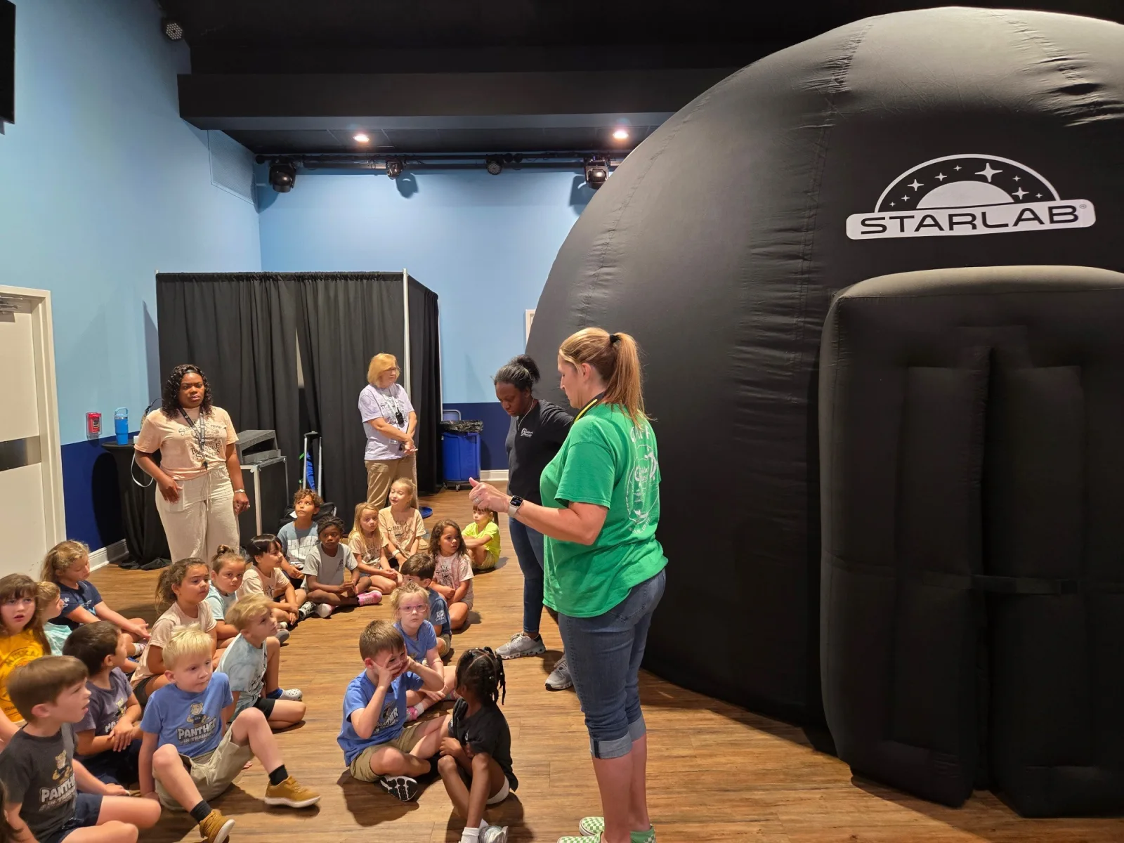 An educator speaks to a group of young students seated on the floor beside a large black inflatable STARLAB planetarium dome