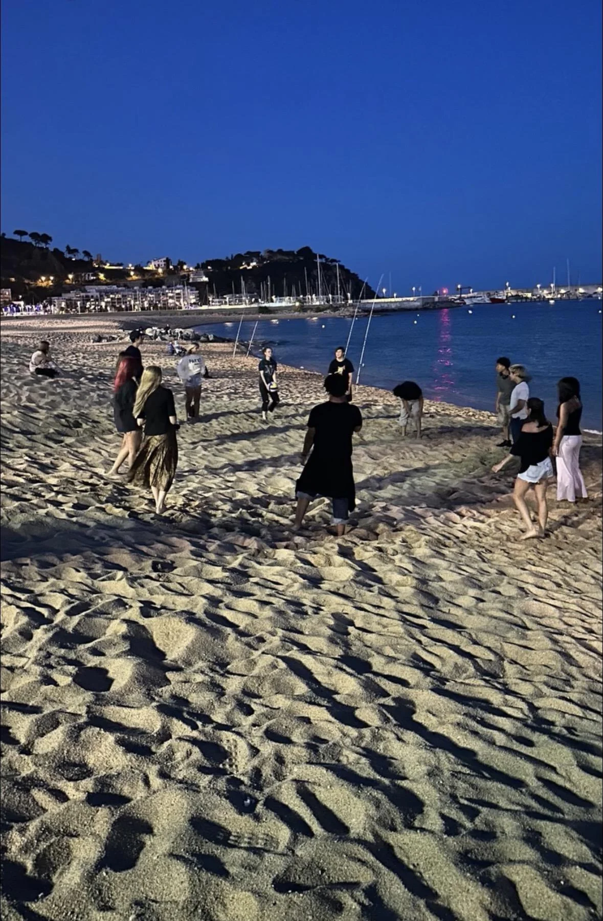 Students walking barefoot on a Spanish beach at dusk, with harbor lights across the bay
