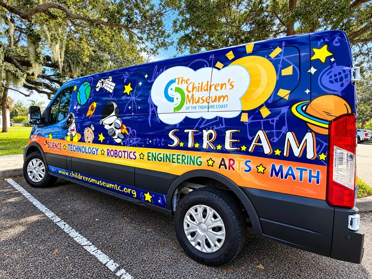 The Children’s Museum of the Treasure Coast Mobile Museum, a blue van wrapped in space art and the words S.T.R.E.A.M., parked under oak trees